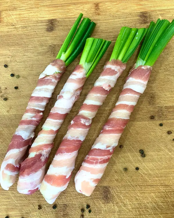 Quattro cipollotti avvolti nella pancetta su un tagliere di legno, con pepe in grani