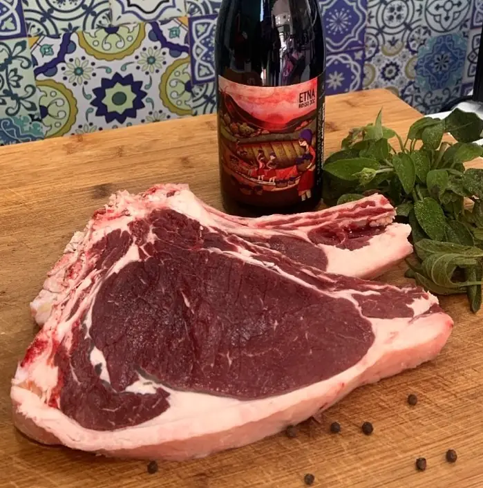 Costata di carne rossa con osso su un tagliere di legno, con salvia, grani di pepe e una bottiglia sullo sfondo