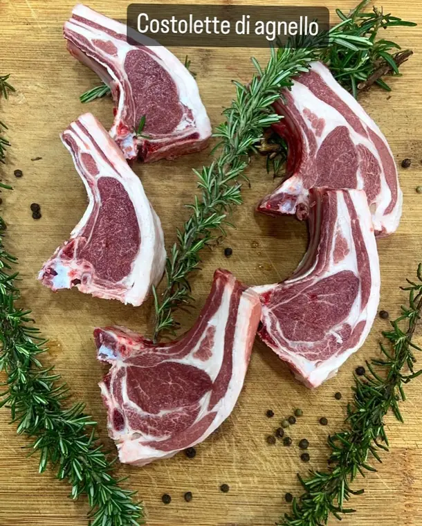 Costolette di agnello crude su un tagliere con rosmarino e pepe nero, con la scritta «Costolette di agnello»