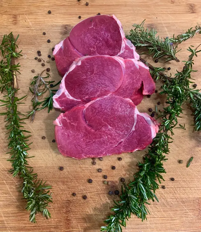 Tre fettine di carne rossa cruda su un tagliere di legno, con rametti di rosmarino e grani di pepe