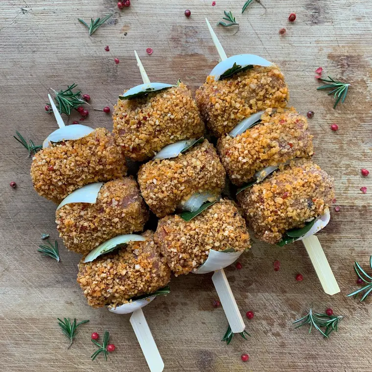 Spiedini di involtini con panatura color arancio, cipolla e foglie di alloro su un tagliere con rosmarino