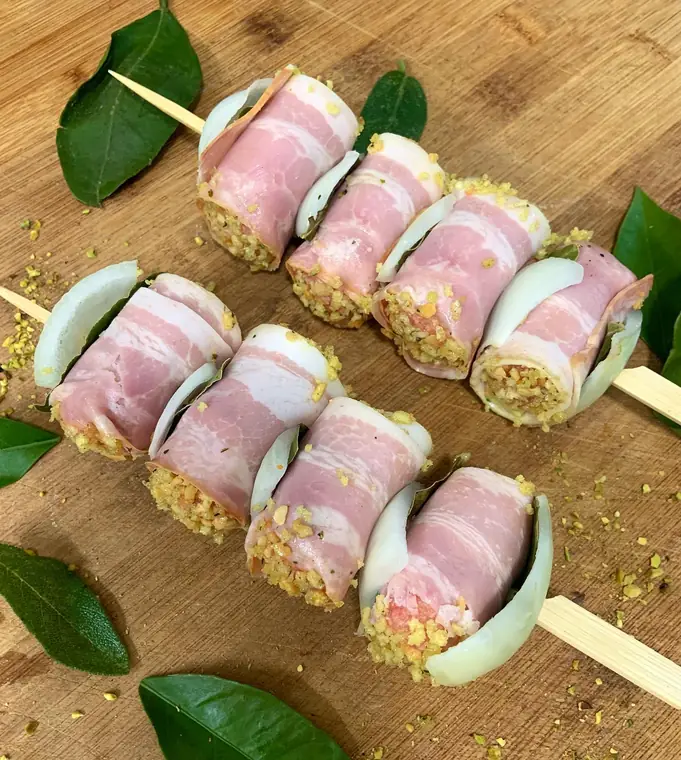 Due spiedini di involtini avvolti nella pancetta, con granella verde, cipolla e foglie di alloro