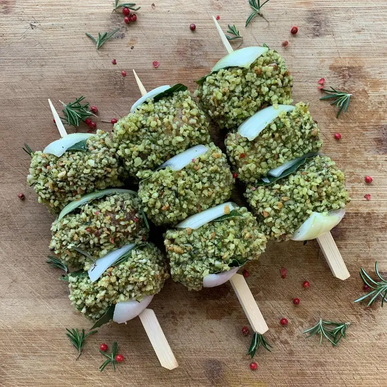 Spiedini di involtini con panatura verde, cipolla e alloro su un tagliere con rosmarino e pepe rosa