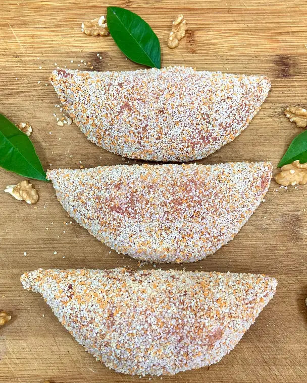Tre preparazioni di carne a mezzaluna con panatura chiara e arancio, tra foglie verdi e noci, su un tagliere
