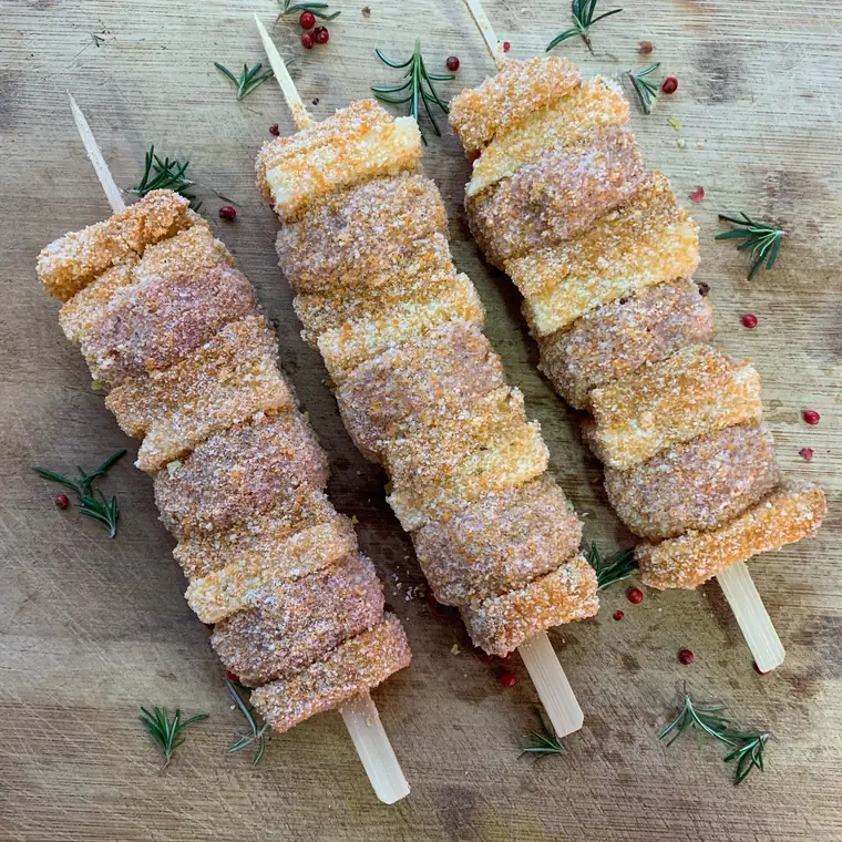 Tre spiedini di carne impanata su un tagliere di legno con rosmarino