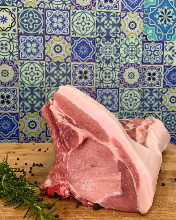 Taglio di carne rosa con osso e cotenna su un tagliere, davanti a una parete di piastrelle colorate, con rosmarino e pepe